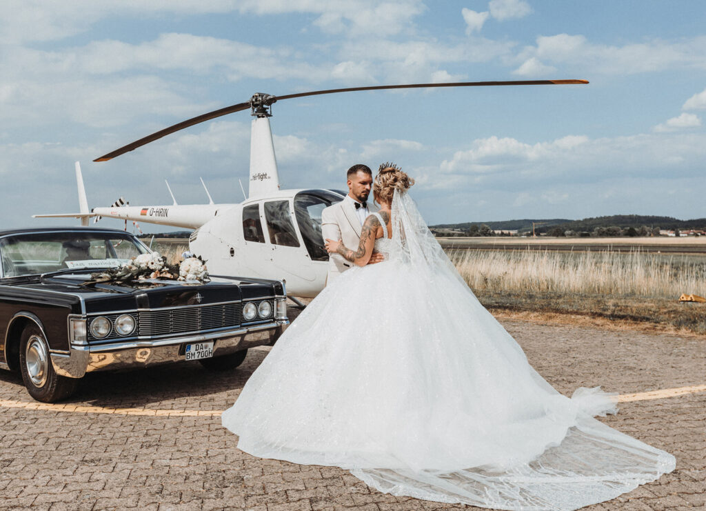 Ein Brautpaar küsst sich vor einem Hubschrauber und einem klassischen Auto auf einem Landeplatz.