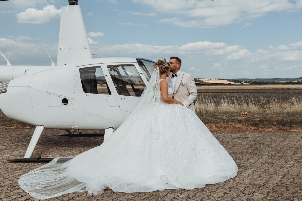 Ein Brautpaar küsst sich neben einem Hubschrauber auf einem offenen Feld.